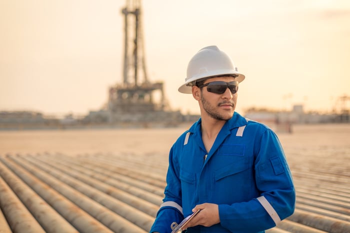 A person wearing personal protective equipment with an oil field and drilling rig in the background.