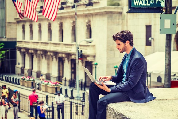 A man sitting on a ledge looking at laptop in front of a street sign reading Wall St.
