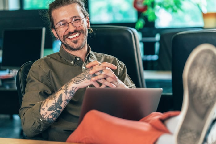 Someone is smiling with his feet up on a table, laptop in lap.