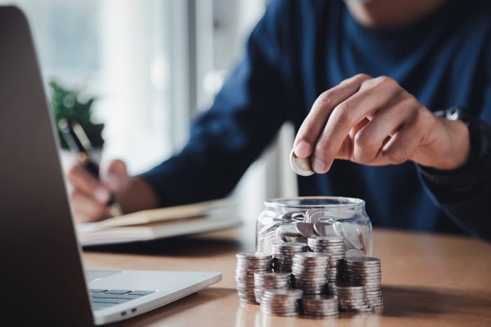 A person sitting at a table in front of a laptop computer and filling a jar with coins. 