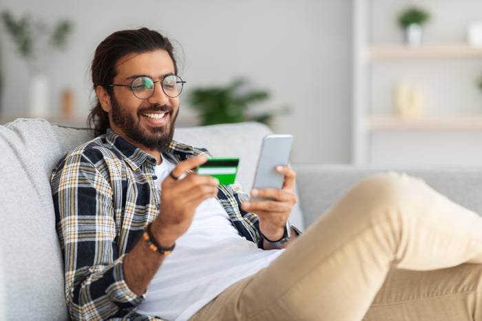 An individual in a living room holds up a credit card and looks at a phone.