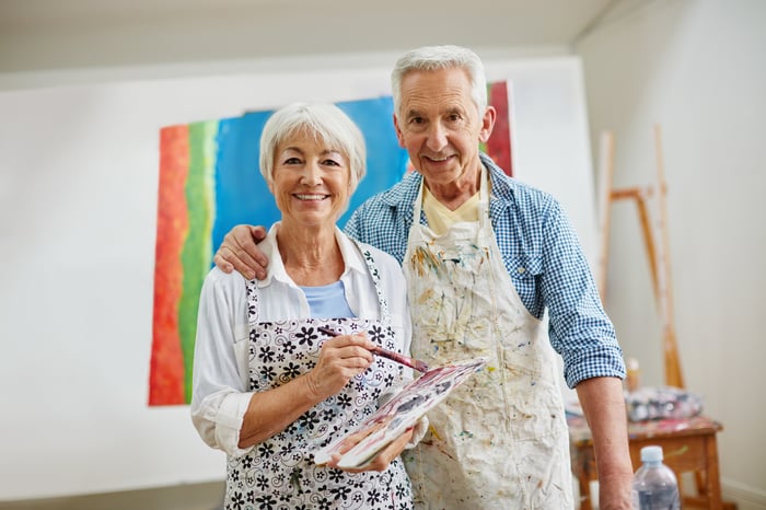 A couple stands in front of a painting with a brush and palette, smiling.