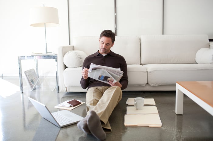 Person sitting against a couch reading a newspaper.