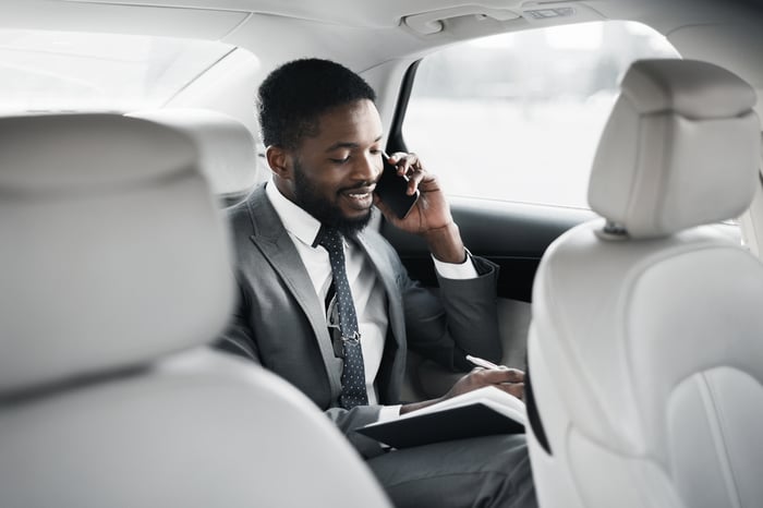 A person sitting in the back of a car talking on the phone while writing notes in a notebook.