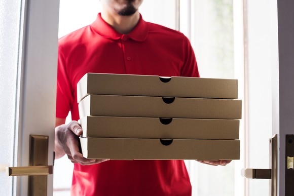 A delivery person drops off four boxes of pizza at a residence, with the picture showing them standing just inside the doorway.