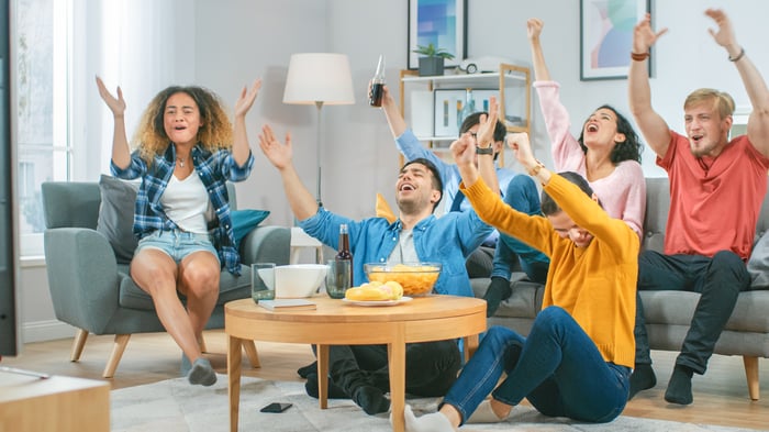 People celebrating while sitting on and around a couch with various snack foods while watching sports on TV.. 