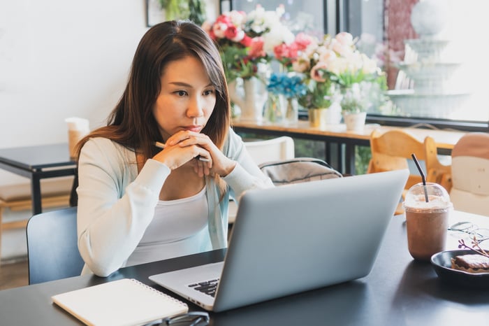 A person sitting at a table and staring intently at a laptop computer with a beverage and pastry on the table.