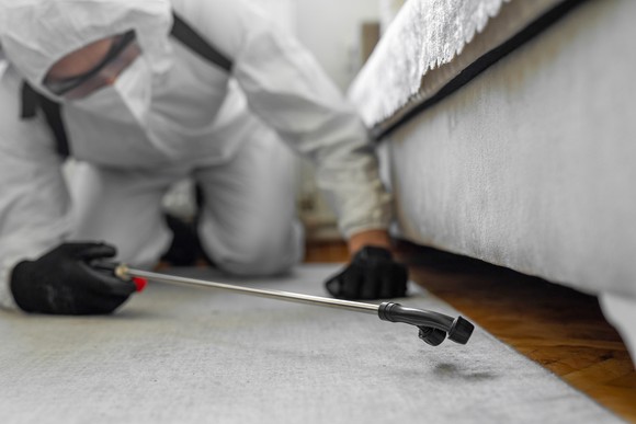 An exterminator dress in white personal protective gear sprays underneath of a couch  in someone's home.