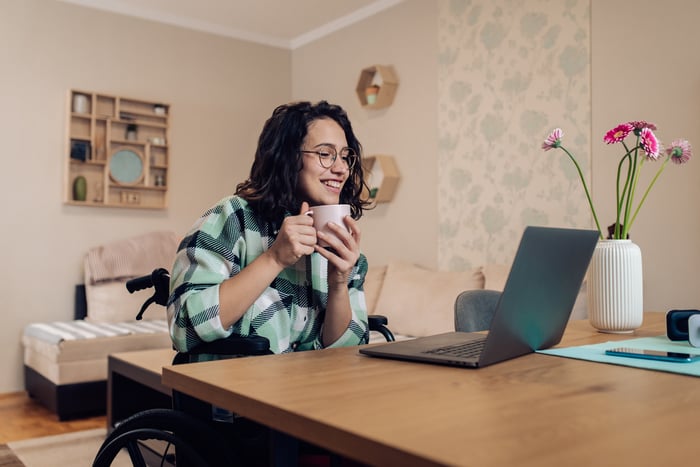 A person in a wheelchair holding a coffee mug while looking at an open laptop at home.