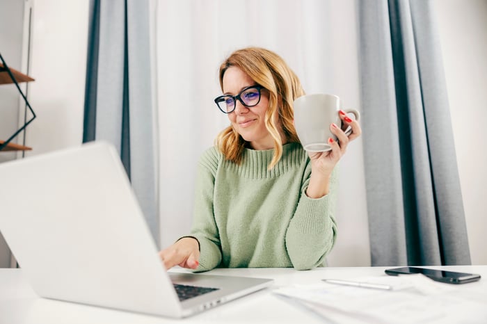 A person smiles while holding a mug and typing on a laptop computer. 
