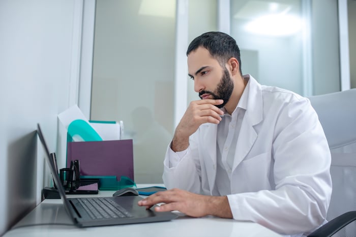 A person in a lab coat sits at a table and looks at a computer. 