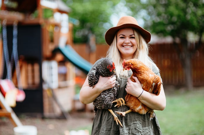 Someone in a hat is smiling and holding two chickens.