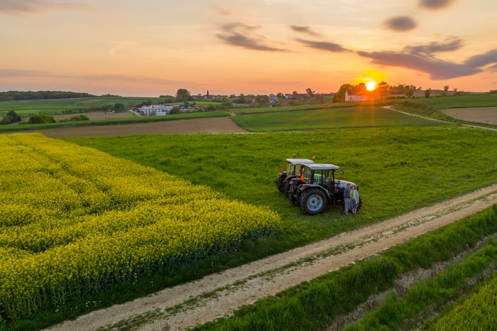 Two people smile while standing by two tractors at sunset on a farm.