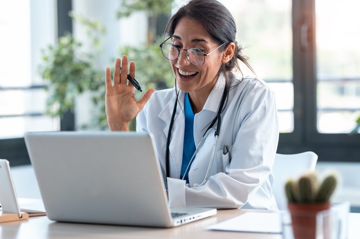 A doctor is smiling and waving at an open laptop.