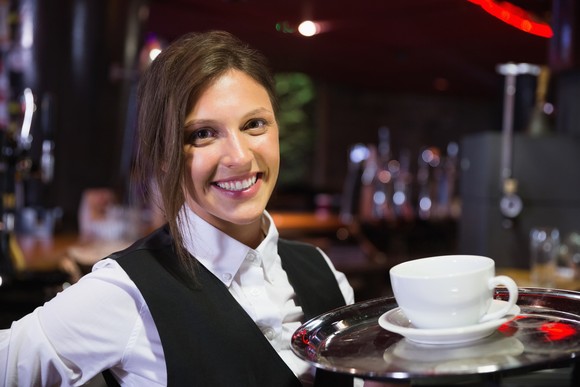 A server is smiling in a restaurant, holding a tray.