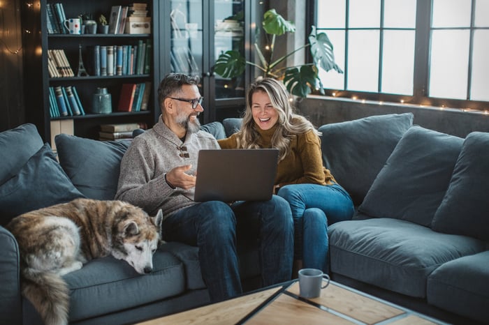 Two people sitting on a couch with a dog and looking at a laptop.