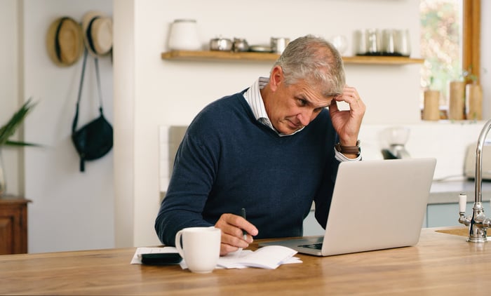 A person scratches their head while sitting at a table and looking at a laptop computer in a concerned manner.