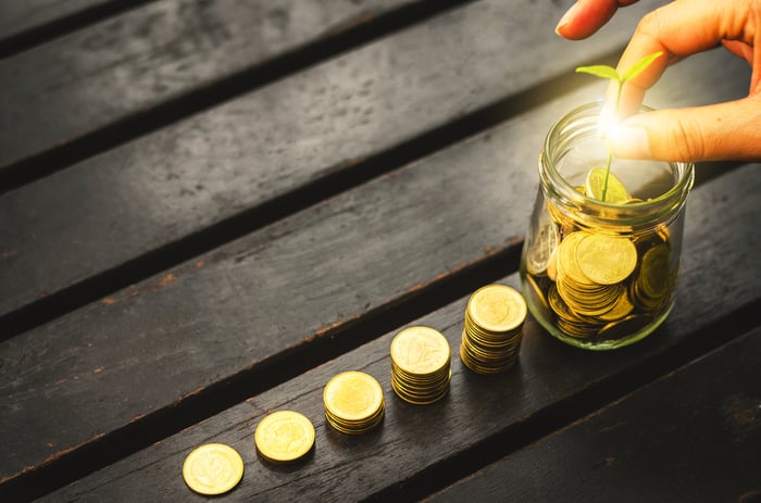 A person touches a glowing sapling in a glass jar of coins next to increasingly taller stacks of coins. 
