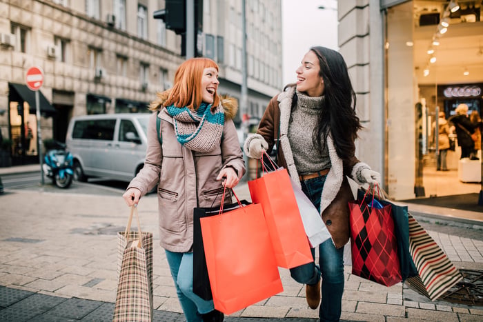 Two people smile while carrying multiple shopping bags and walking down the street.