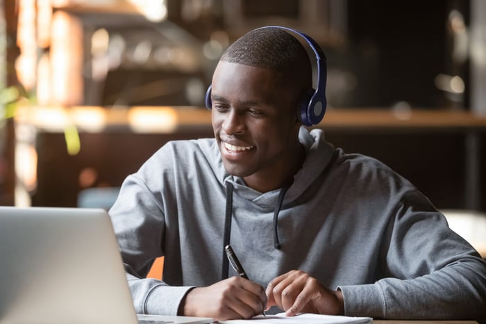 A person wearing headphones smiles and holds a pen to a pad of paper while sitting in front of a laptop computer.