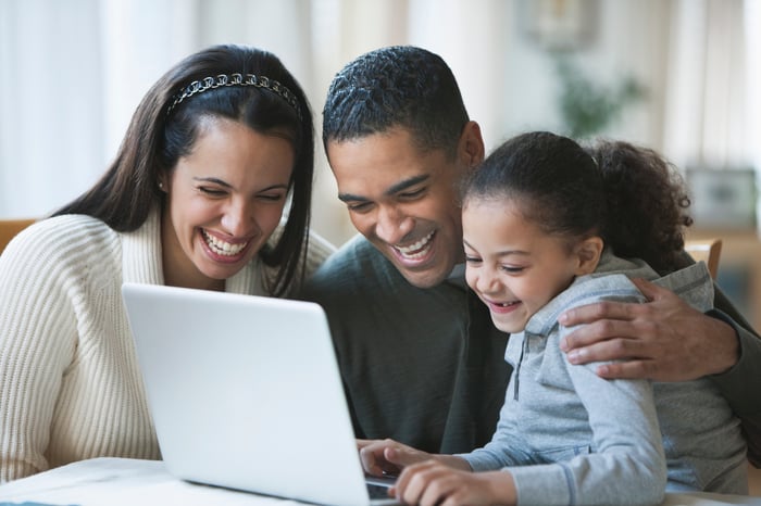 Two adults and a child smile while looking at something on a laptop at home.