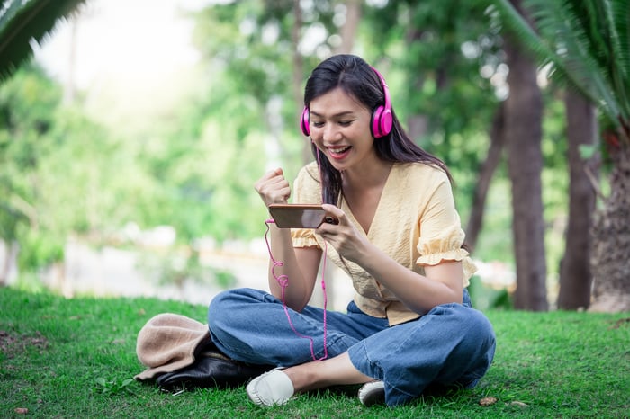 A person with pink headphones looking at a phone and cheering as they sit on a lawn.