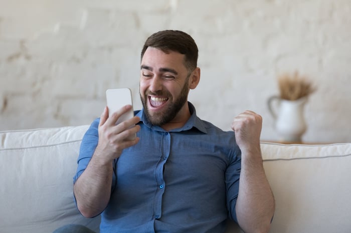 Person sitting on an off-white couch holding a rectangular object in hand with a vibrant facial expression.