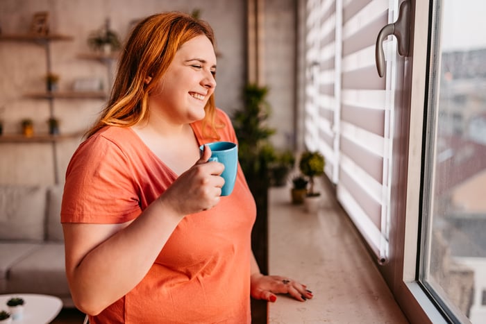 A person smiles while holding a mug and looking out a window.