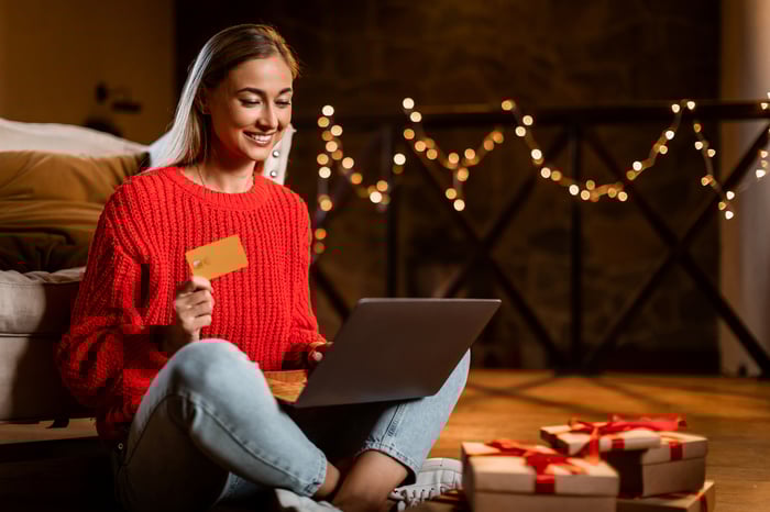 A person holding a payment card and smiling while sitting on the ground with a laptop computer next to presents. 