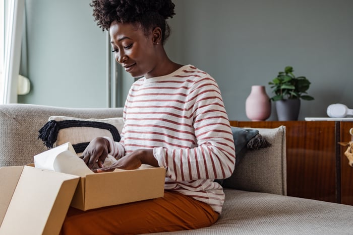 A person smiles while sitting on a couch and packing items in a cardboard box.