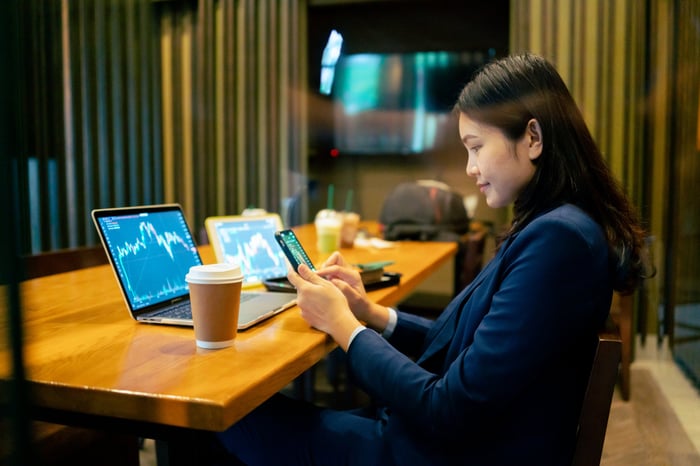 An investor checks a portfolio on a phone and a laptop computer in a coffee shop.