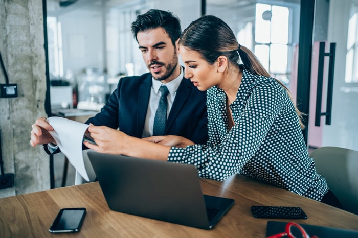 Two investors in an office look at documents near a laptop.