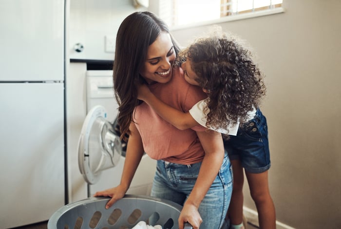 An adult holding a laundry basket smiles at a child wrapped around the adult's shoulders.