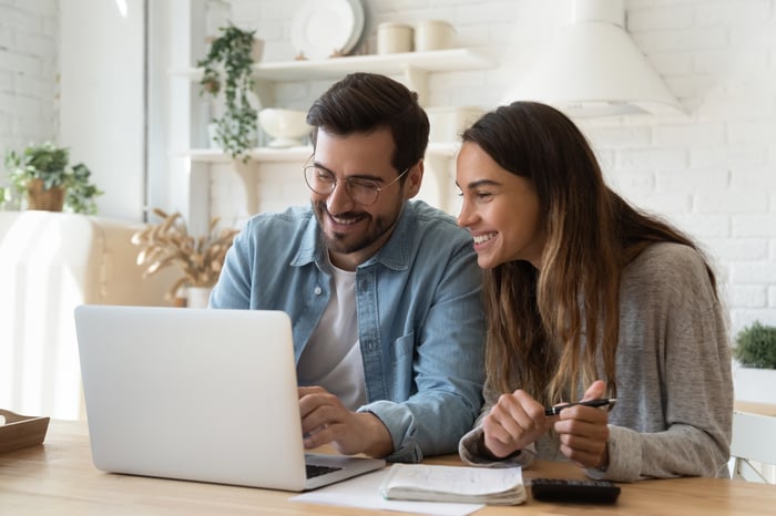 Two investors smile while looking at something on a laptop in their living room.