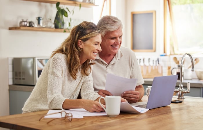 Two people smile while sitting at a table holding pieces of paper and looking at a laptop computer. 