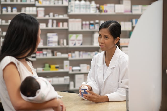 A pharmacist holding a prescription while speaking with a customer who has an infant in their arms.