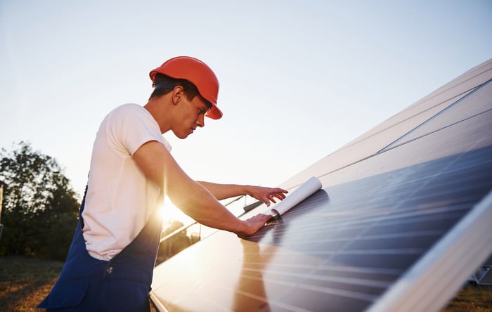 A worker wearing a hard hat is looking at papers on a solar panel.