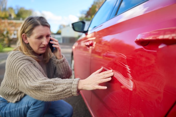 A person is on the phone while looking at a scratch on their car.