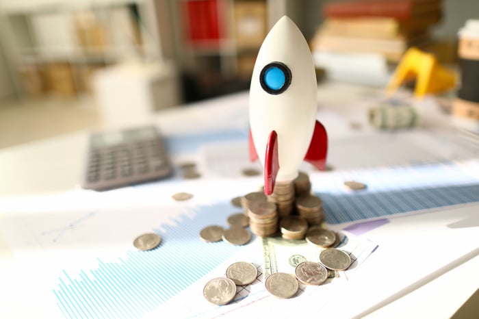 A toy rocket set for launch atop stacks of coins and paperwork displaying financial data.