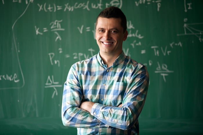 A teacher is standing in front of a blackboard with arms crossed, smiling.