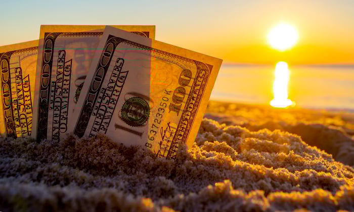 Three fanned one hundred dollar bills partially buried standing up in the sand, with the sun rising on the horizon.