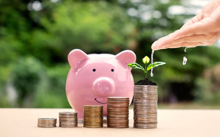 Water droplets fall from a person’s hand onto a plant sprouting from stacks of coins with a pink piggy bank in the background.