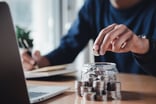 A person sitting at a table in-front of a computer putting coins into a glass jar.
