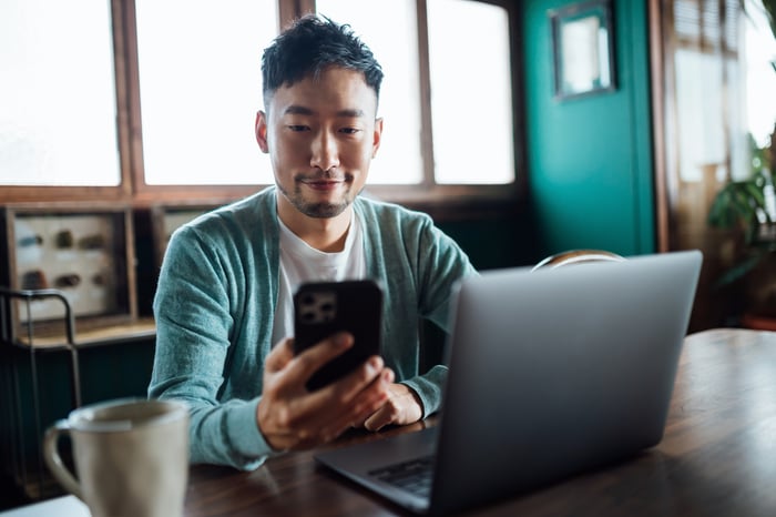 A person smiles while sitting at a table in front of a laptop computer and looking at their cell phone.