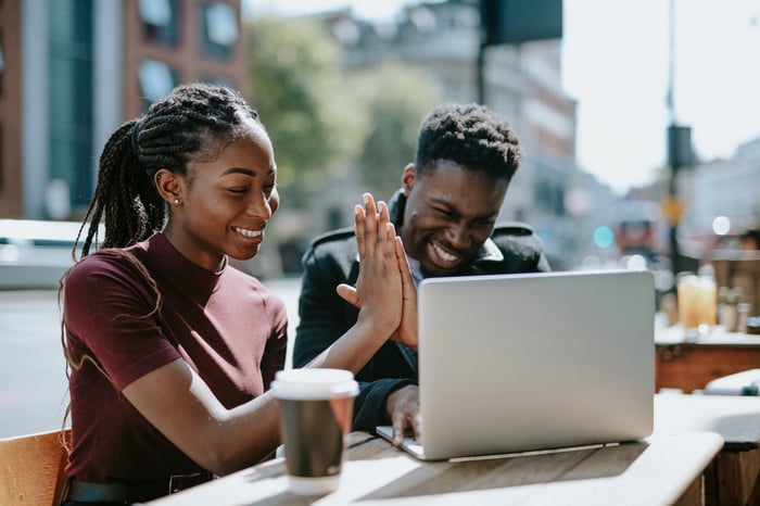 Two people smile and high-five while sitting at a table in a public setting in-front of a laptop computer.