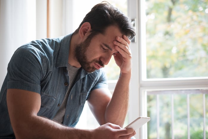 A person puts their hand on their forehead while sitting down and looking at a mobile phone in a concerned manner.
