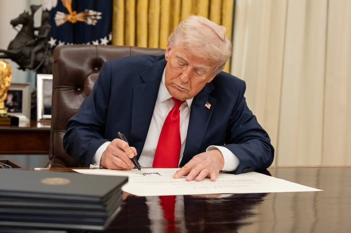 President Donald Trump signing a piece of paper while sitting at a desk.