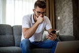 A person sitting on a couch looking at their phone with their first covering their mouth.
