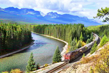 A train passing by a river in a forest with mountains in the background.