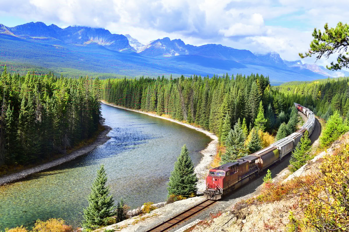 A train passing by a river in a forest with mountains in the background.